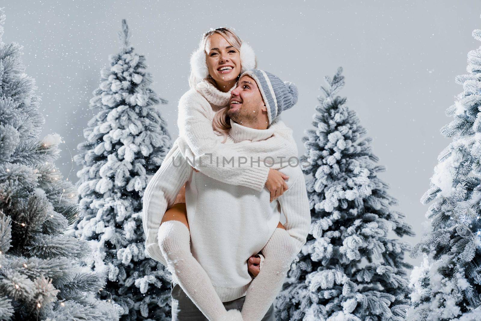Couple kiss and hug. Man holds girl near christmas trees in winter day. New year celebration. People weared wearing fur headphones, hats, white sweaters.