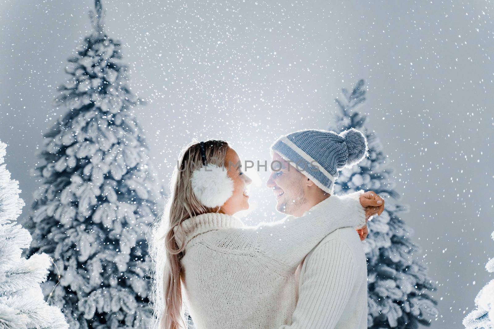 Happy young couple hugs and smiles and snow falls near christmas trees at the eve of new year celebration in winter day. Smiley man and woman weared white pullovers love each other. by Rabizo