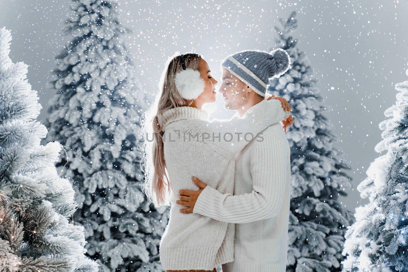 Falling snow and kisses. Happy young couple close-up hugs and kiss near christmas trees at the eve of new year celebration in winter day. Smiley man and woman weared white pullovers love each other. by Rabizo