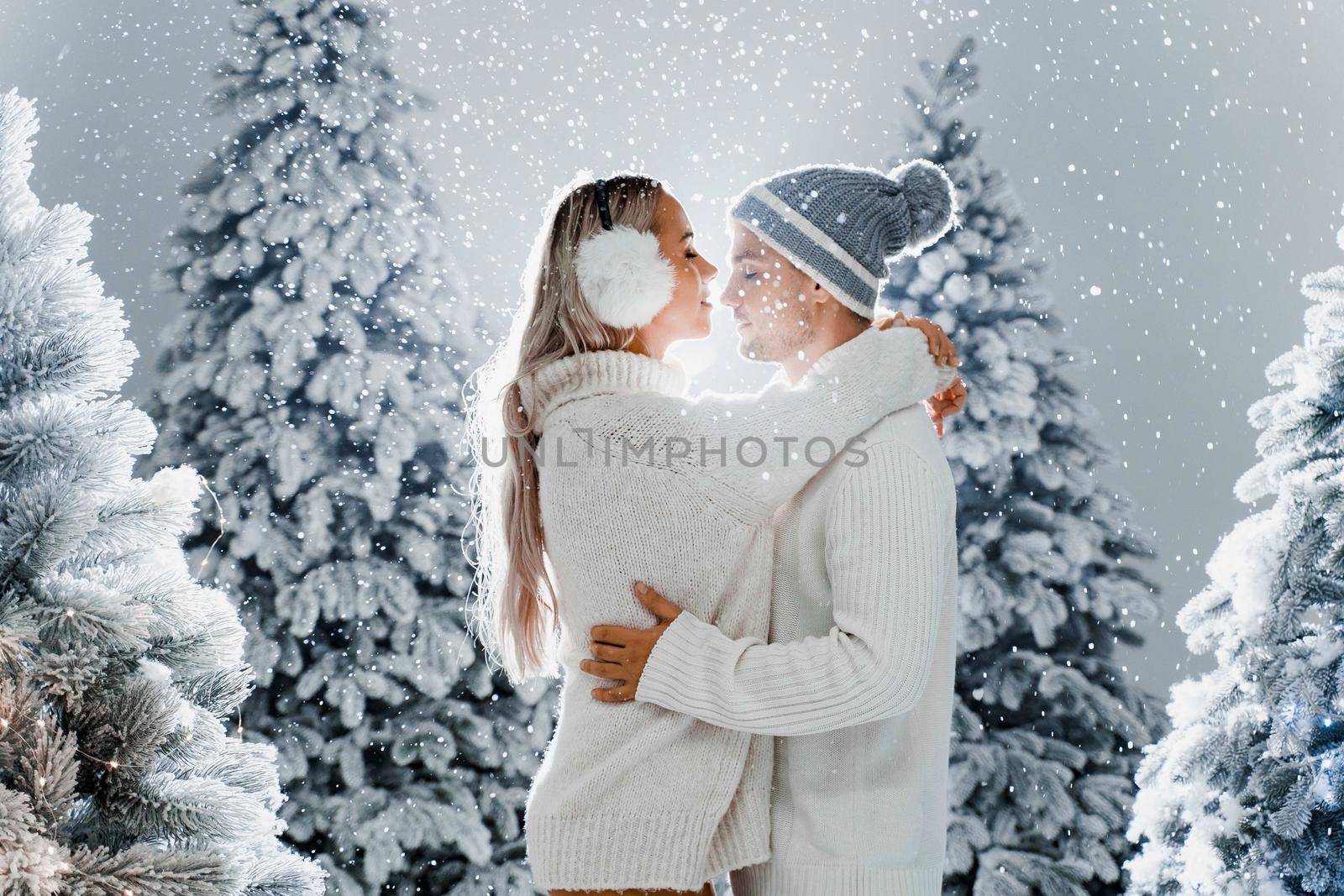 Falling snow and kisses. Happy young couple close-up hugs and kiss near christmas trees at the eve of new year celebration in winter day. Smiley man and woman weared white pullovers love each other