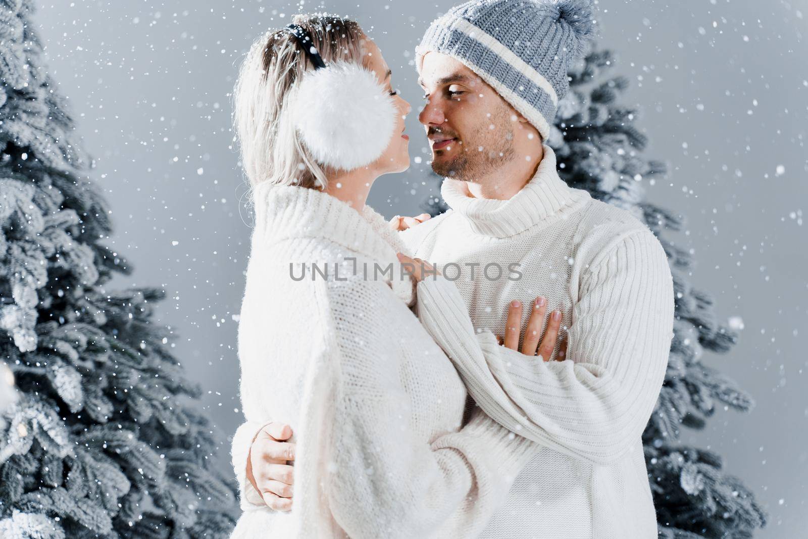 Couple kissing while snow falls near christmass trees. Winter holidays. Love story of young couple weared white pullovers. Happy man and young woman hug each other by Rabizo