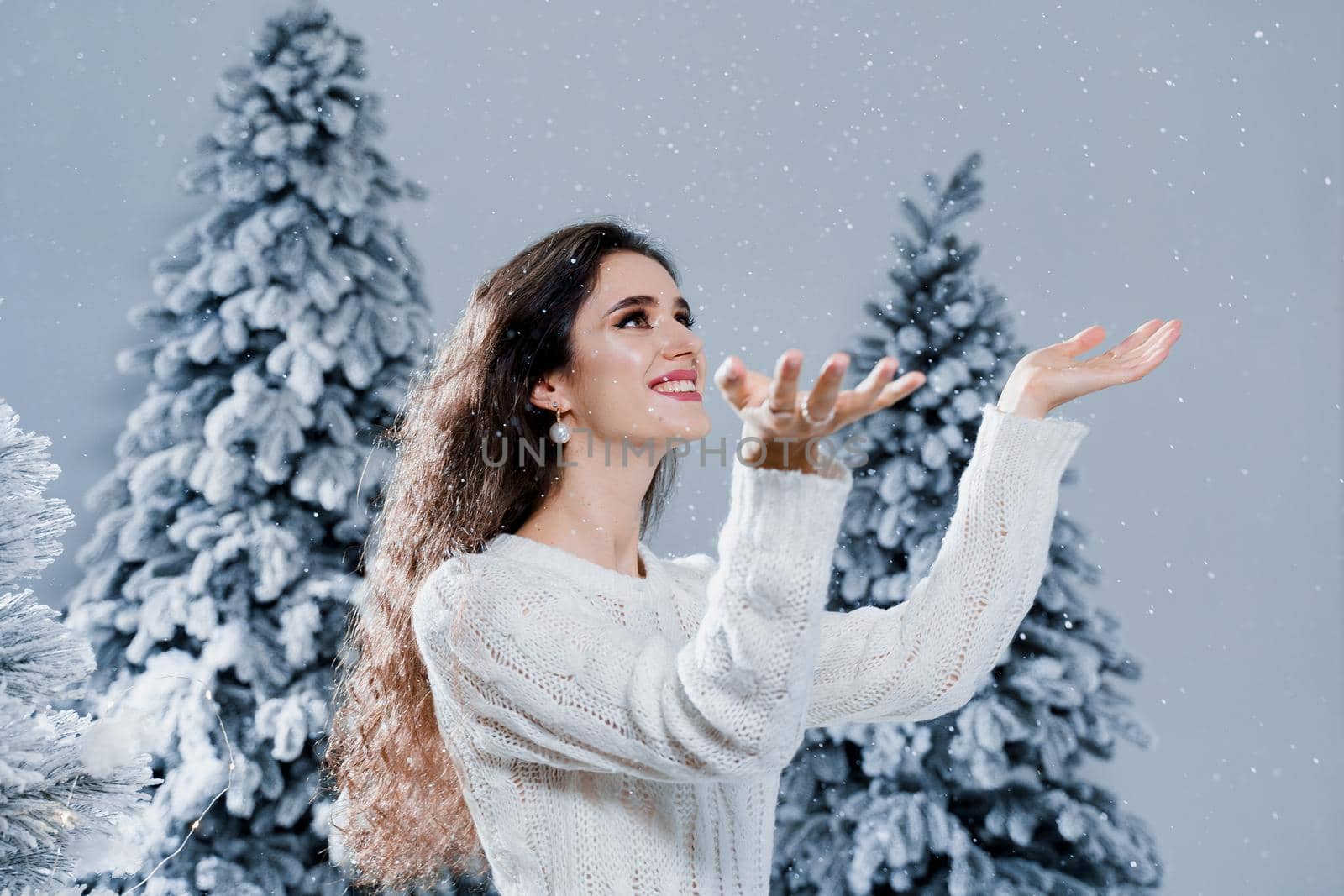 Happy girl smiles and touches falling snow at eve new year. Young woman near big snowy spruces in winter day before christmas celebration