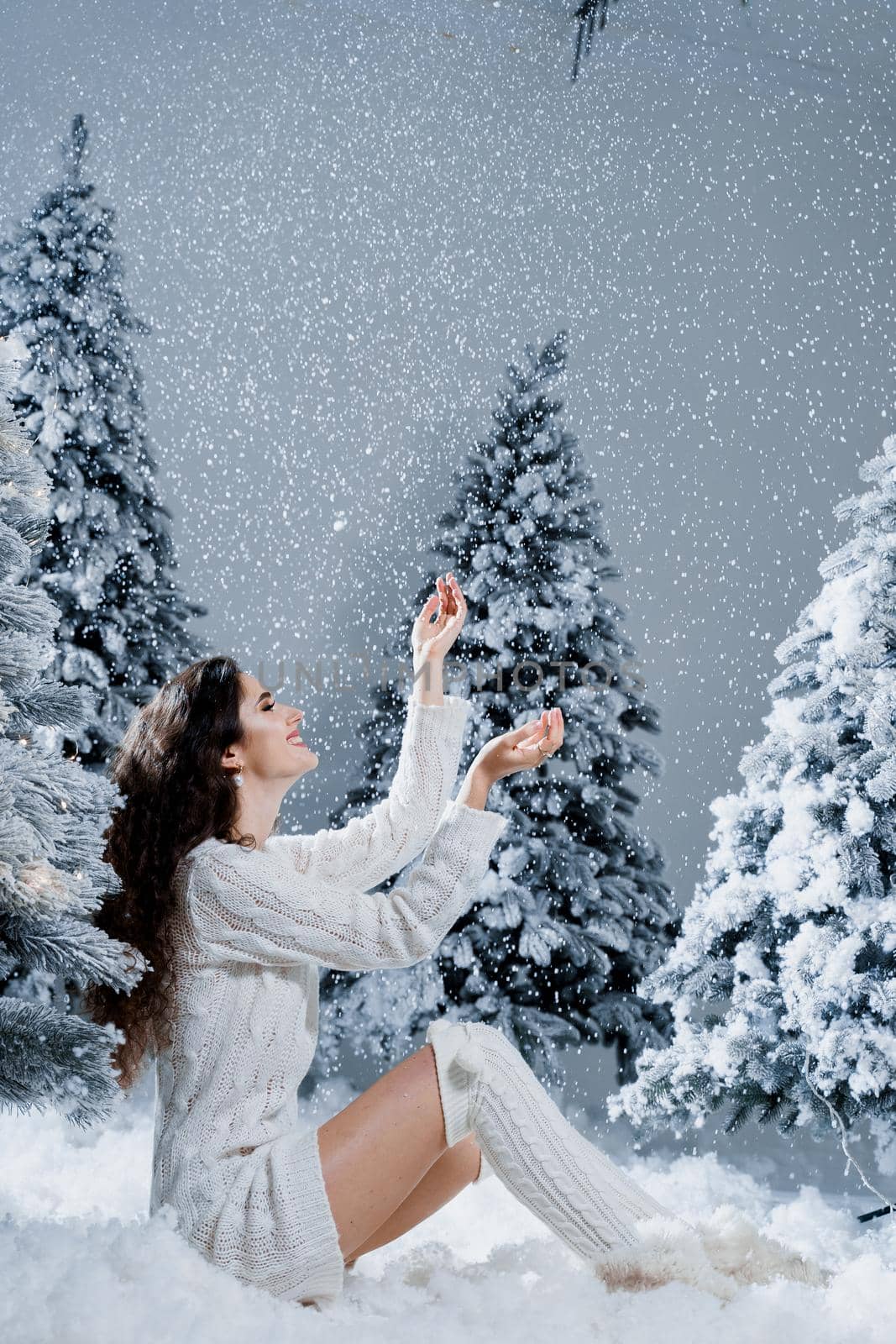 Happy girl smiles and touches falling snow at eve new year. Young woman near big snowy spruces in winter day before christmas celebration. by Rabizo