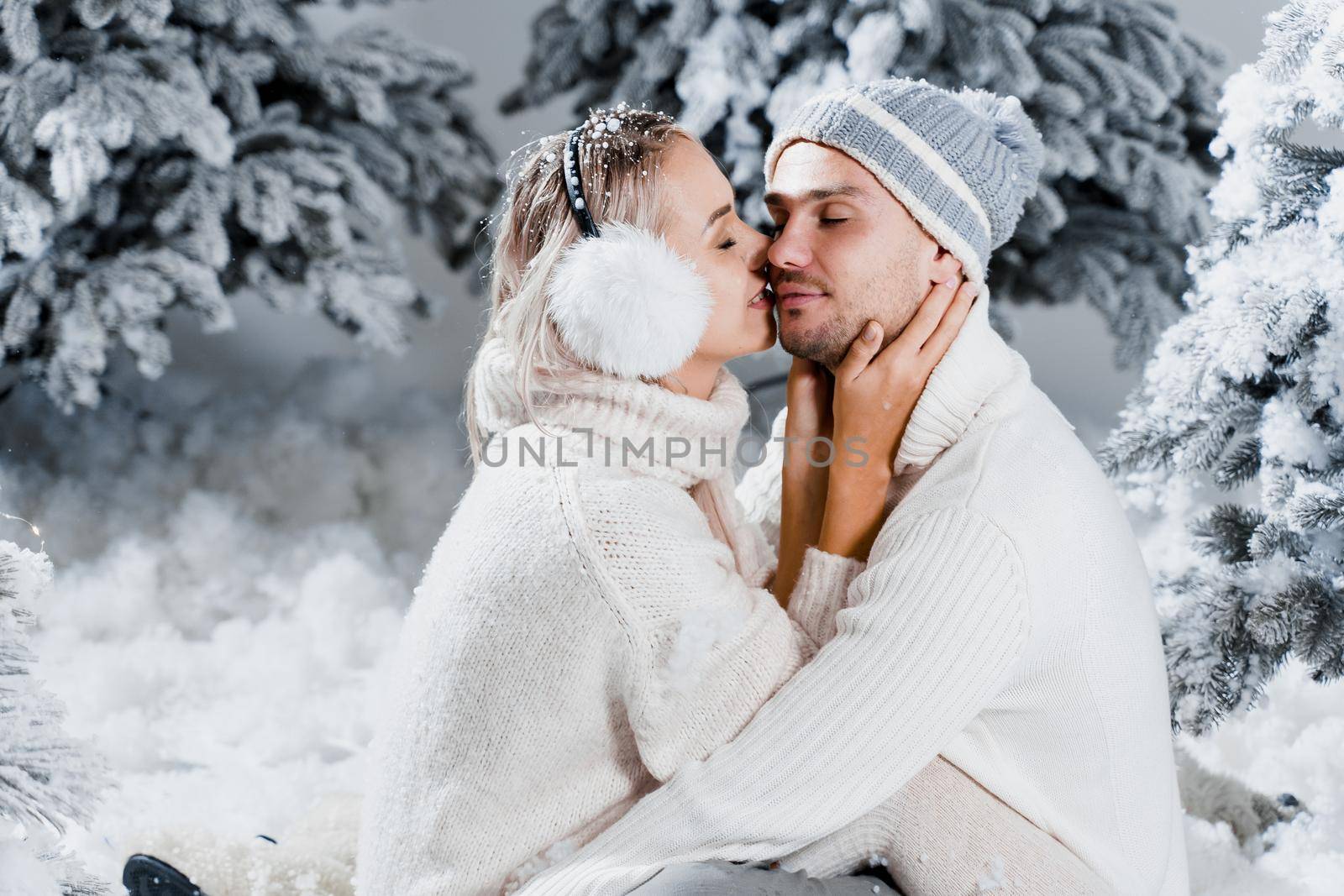 Young woman kissing her lovely man in cheek. Winter holidays. Love story of young couple weared white pullovers. Happy man and young woman hug each other.