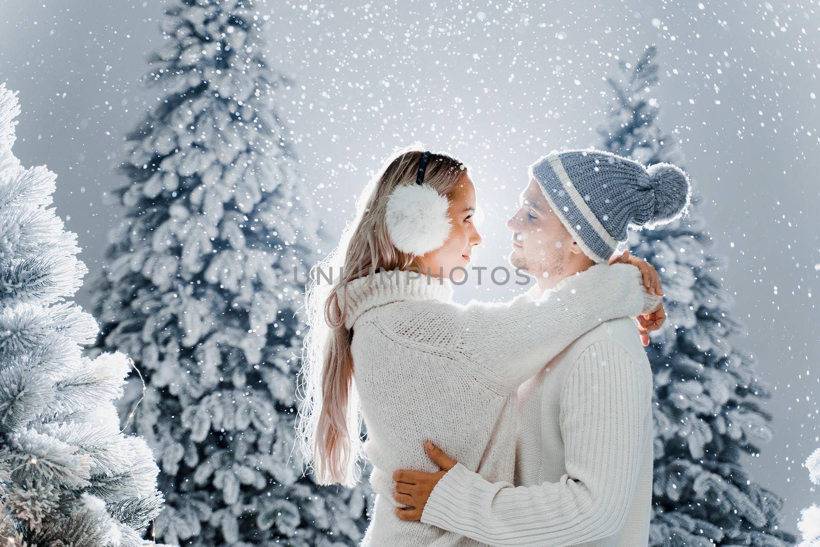 Couple kissing while snow falls near christmass trees. Winter holidays. Love story of young couple weared white pullovers. Happy man and young woman hug each other.