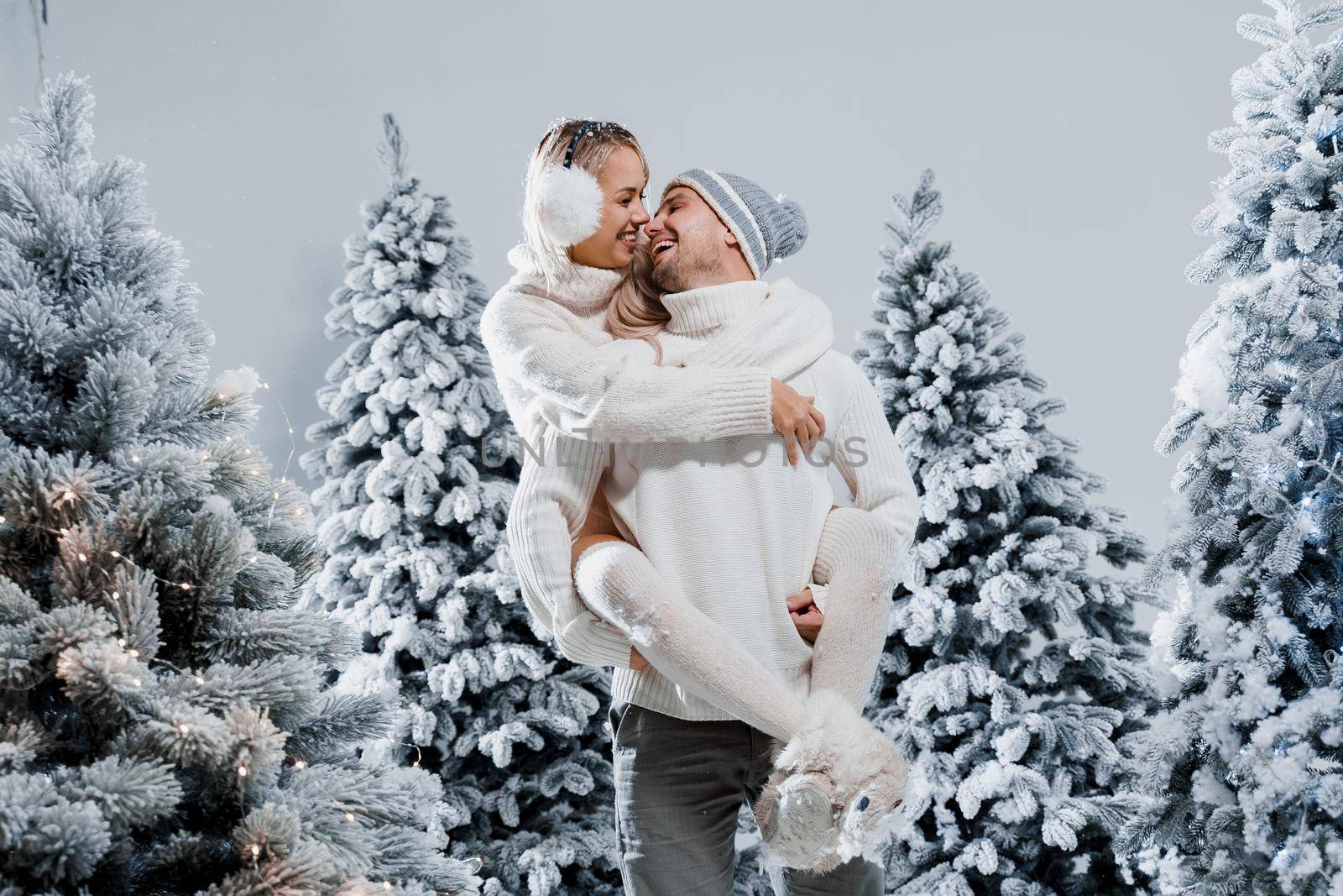 Couple couple laughing and having fun while snow falls near christmass trees. Winter holidays. Love story of young couple weared white pullovers. Happy man and young woman hug each other by Rabizo