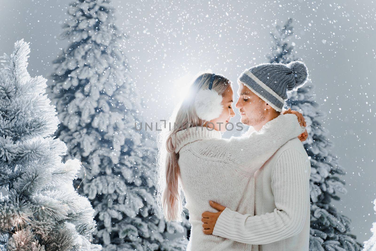 Couple kissing while snow falls near christmass trees. Winter holidays. Love story of young couple weared white pullovers. Happy man and young woman hug each other by Rabizo