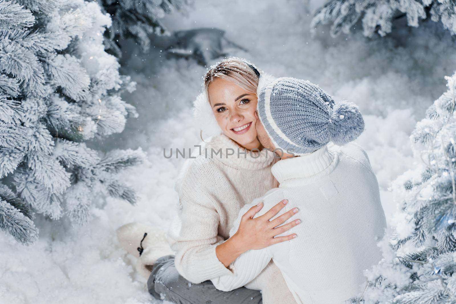 Handsome man is kissing his lovely girl in cheek. Winter holidays. Love story of young couple weared white pullovers. Happy man and young woman hug each other by Rabizo