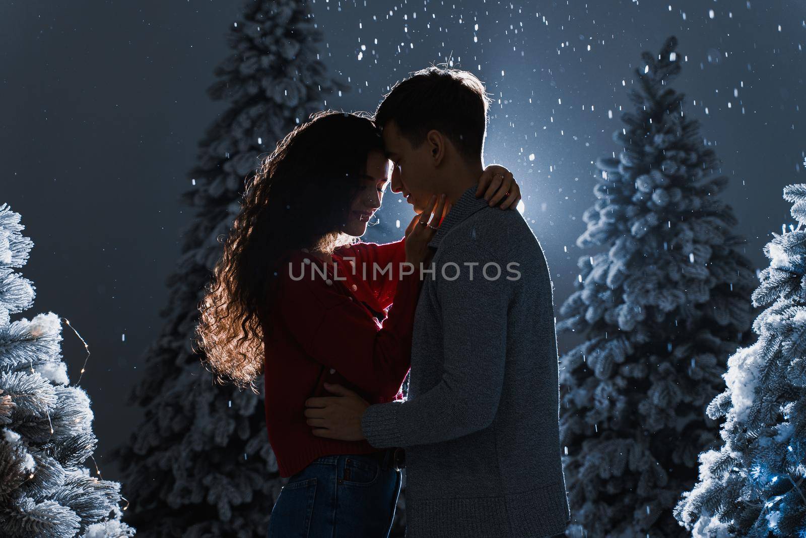 Falling snow and kisses with falling snow on dark blue background. Happy young couple hug and kiss near christmas trees at the eve of new year celebration in winter day. by Rabizo