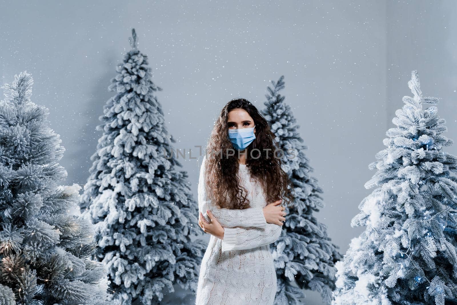 New year celebration at covid-19 coronavirus quarantine period. Happy girl in medical mask with falling snow stays at home. Social distance. Winter holidays in snowy day