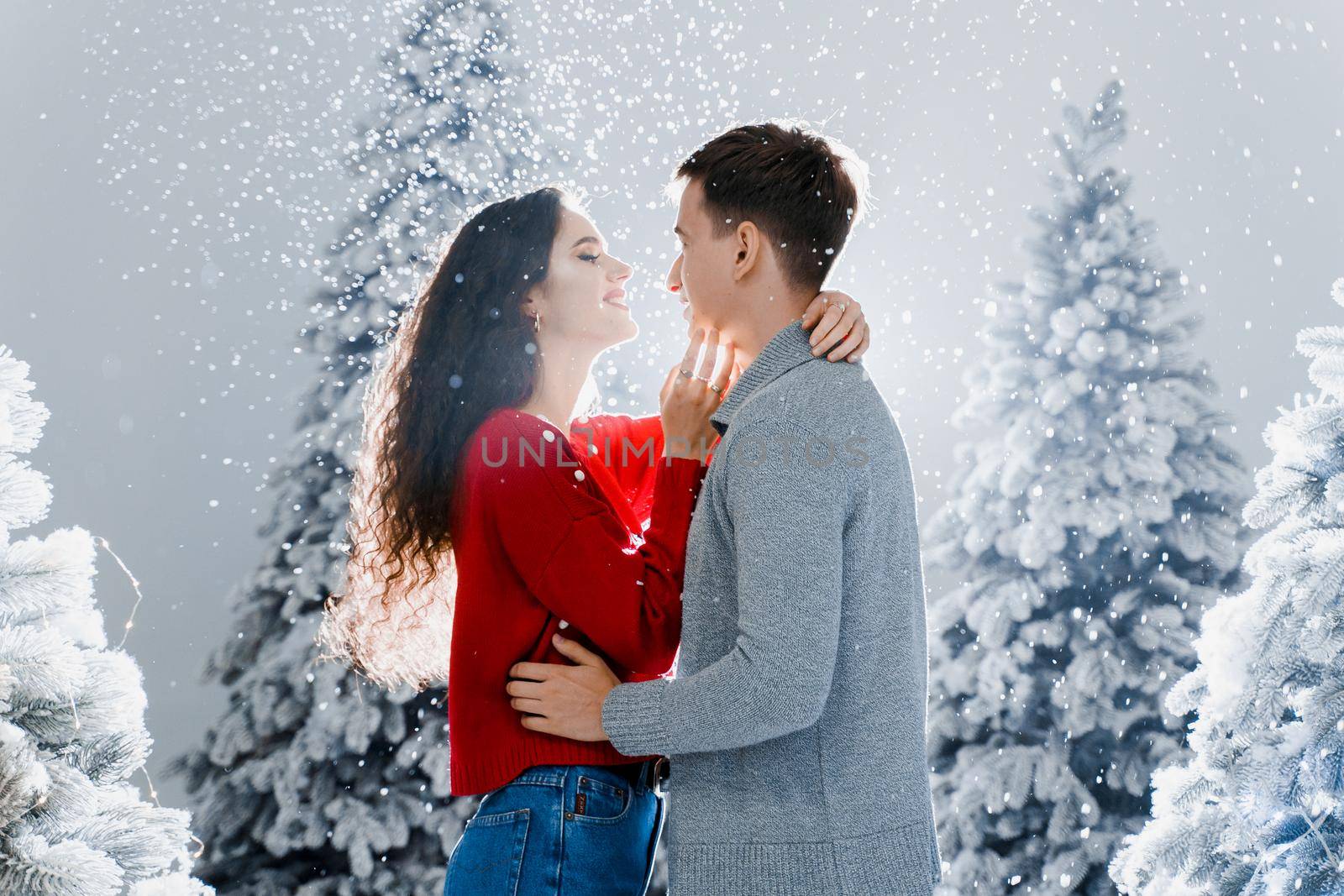 Happy young couple hugs and kiss near christmas trees at the eve of new year celebration in winter day. Smiley man and woman love each other. Falling snow and kisses by Rabizo