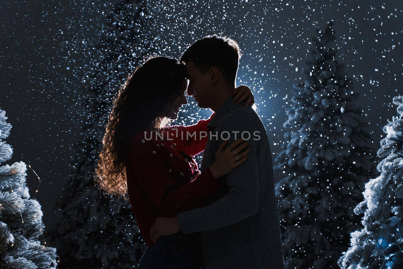 Falling snow and kisses with falling snow on dark blue background. Happy young couple hug and kiss near christmas trees at the eve of new year celebration in winter day