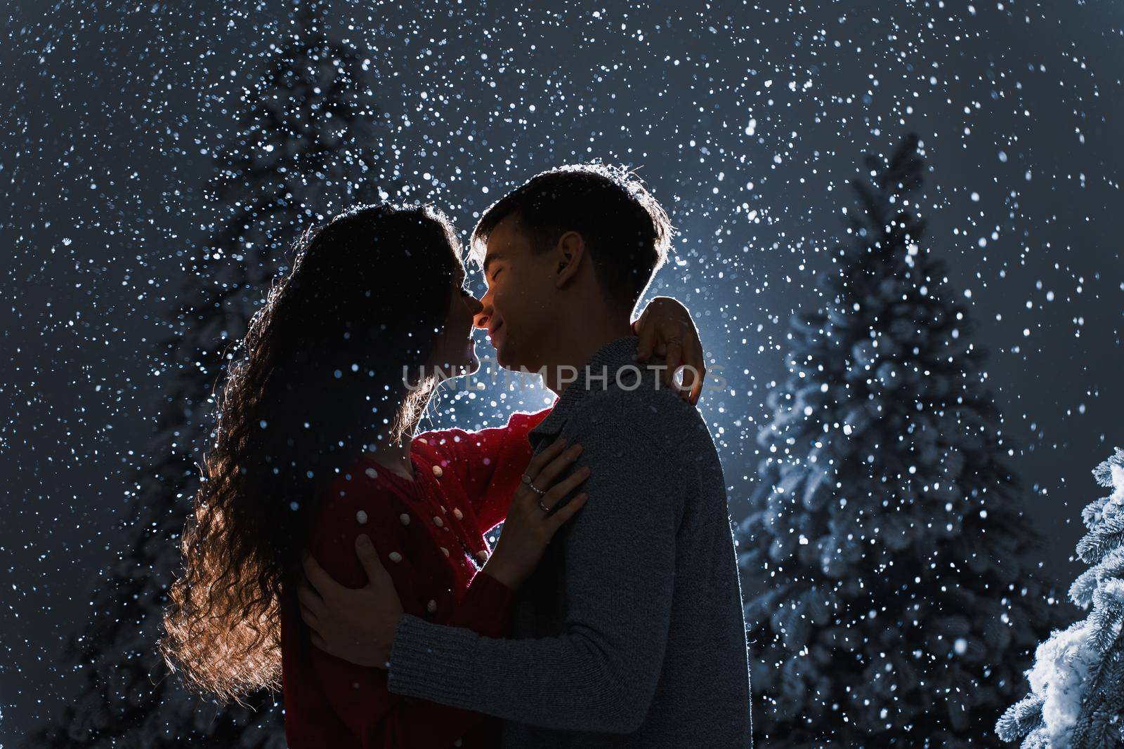 Falling snow and kisses with falling snow on dark blue background. Happy young couple hug and kiss near christmas trees at the eve of new year celebration in winter day