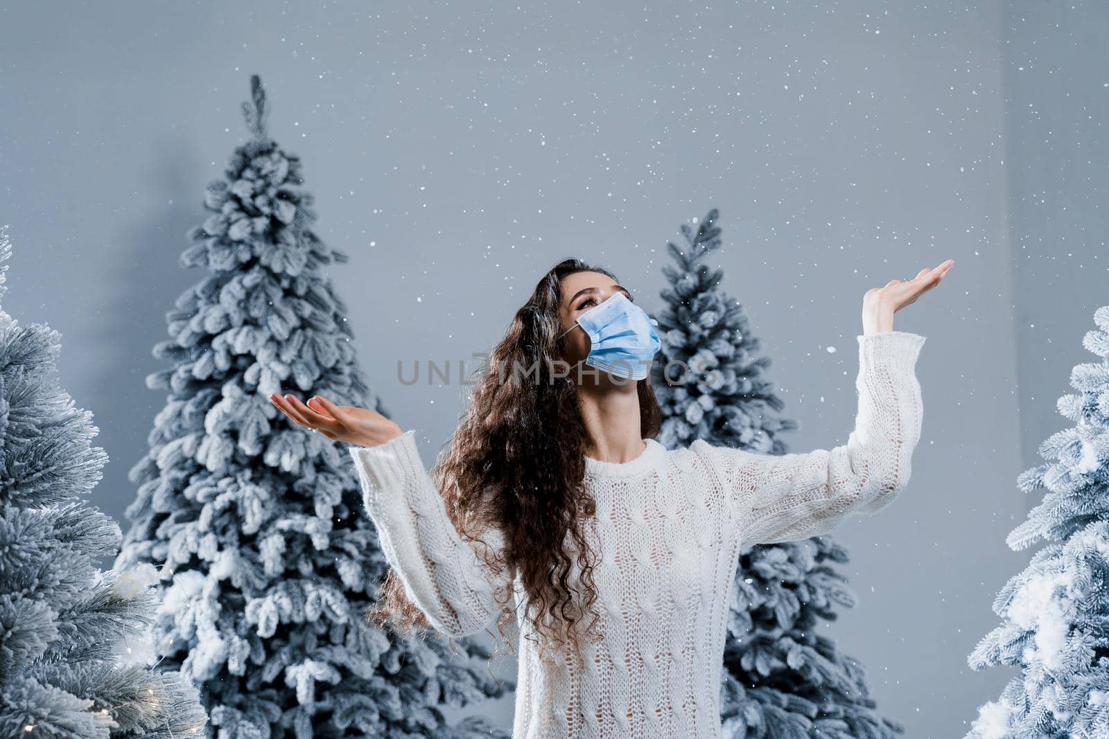 New year celebration at covid-19 coronavirus quarantine period. Happy girl in medical mask with falling snow stays at home. Social distance. Winter holidays in snowy day. by Rabizo