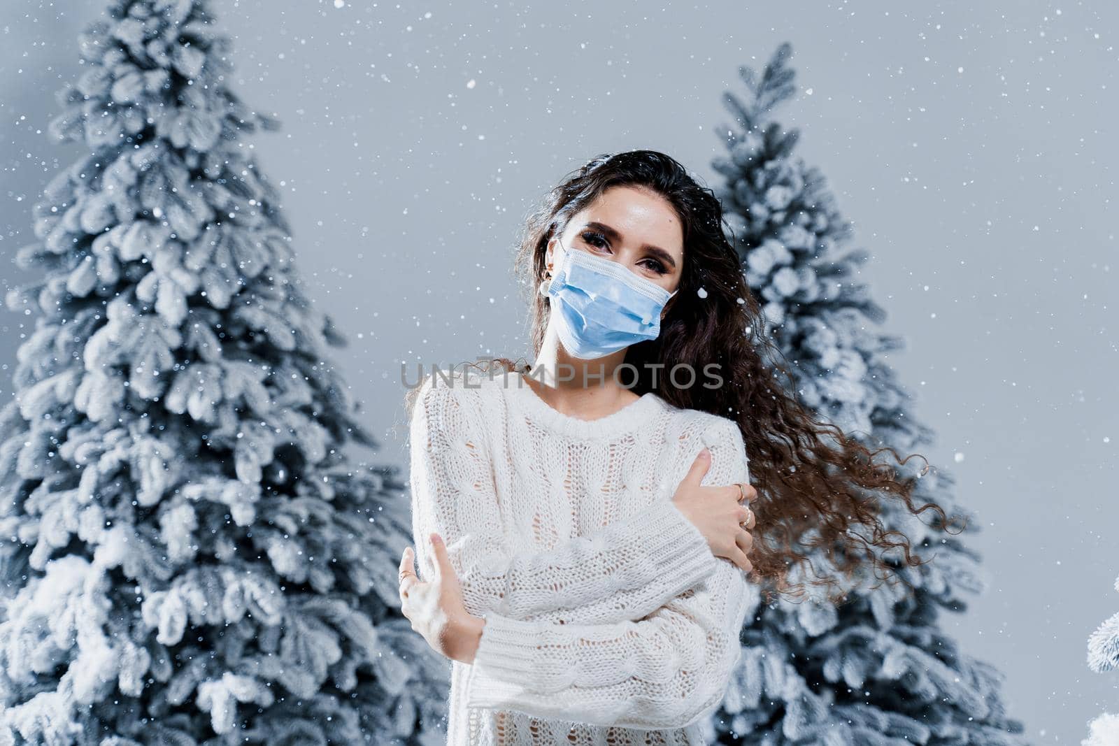 New year celebration at covid-19 coronavirus quarantine period. Happy girl in medical mask with falling snow stays at home. Social distance. Winter holidays in snowy day