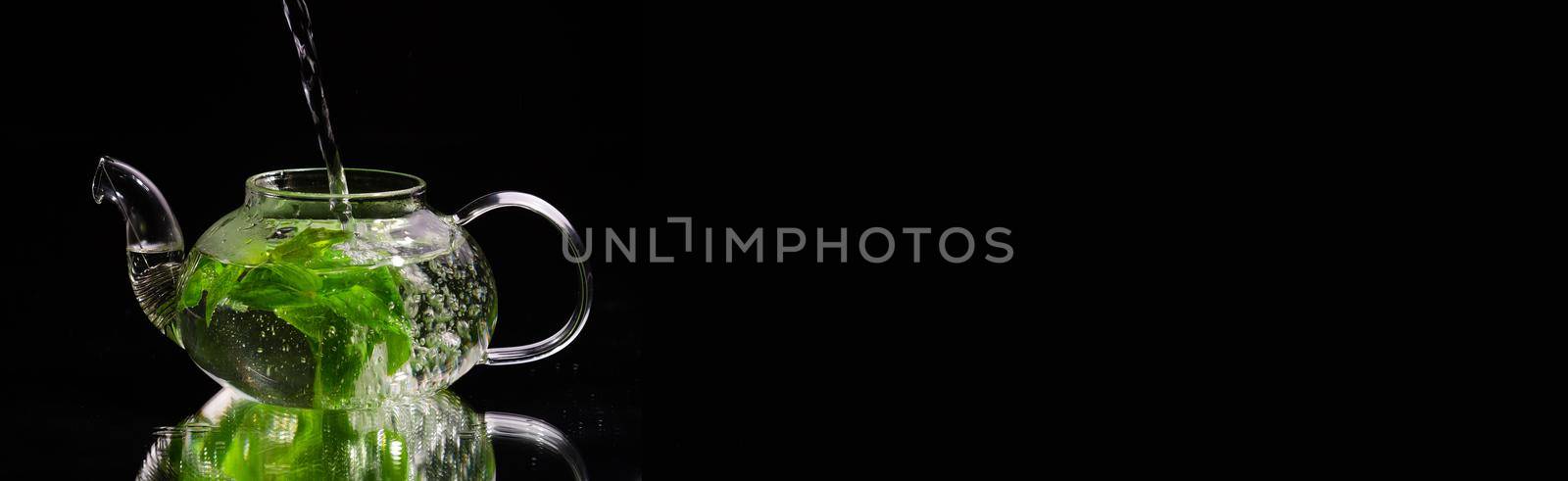 Pouring hot water into a glass teapot on a black background, green mint tea. Herbal tea and healthy drink concept