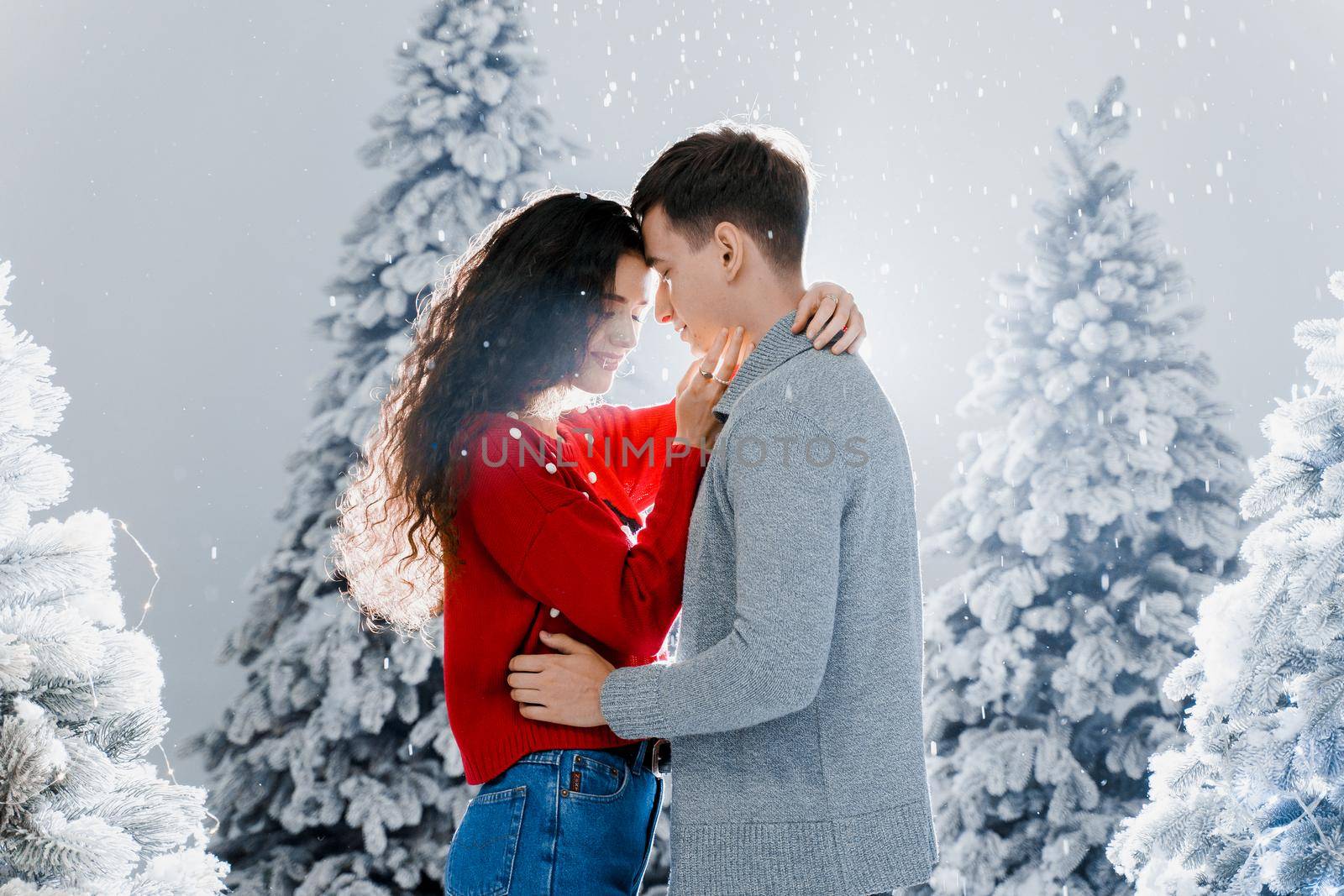 Happy young couple hugs and kiss near christmas trees at the eve of new year celebration in winter day. Smiley man and woman love each other. Falling snow and kisses.