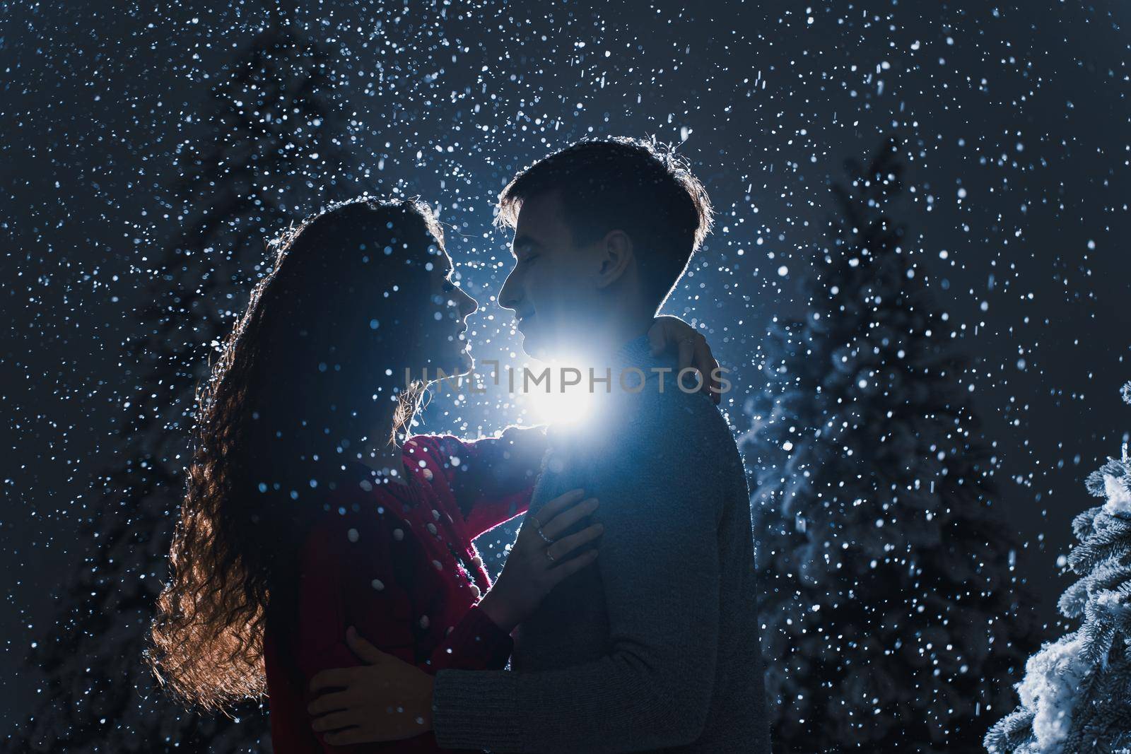 Falling snow and kisses with falling snow on dark blue background. Happy young couple hug and kiss near christmas trees at the eve of new year celebration in winter day. by Rabizo