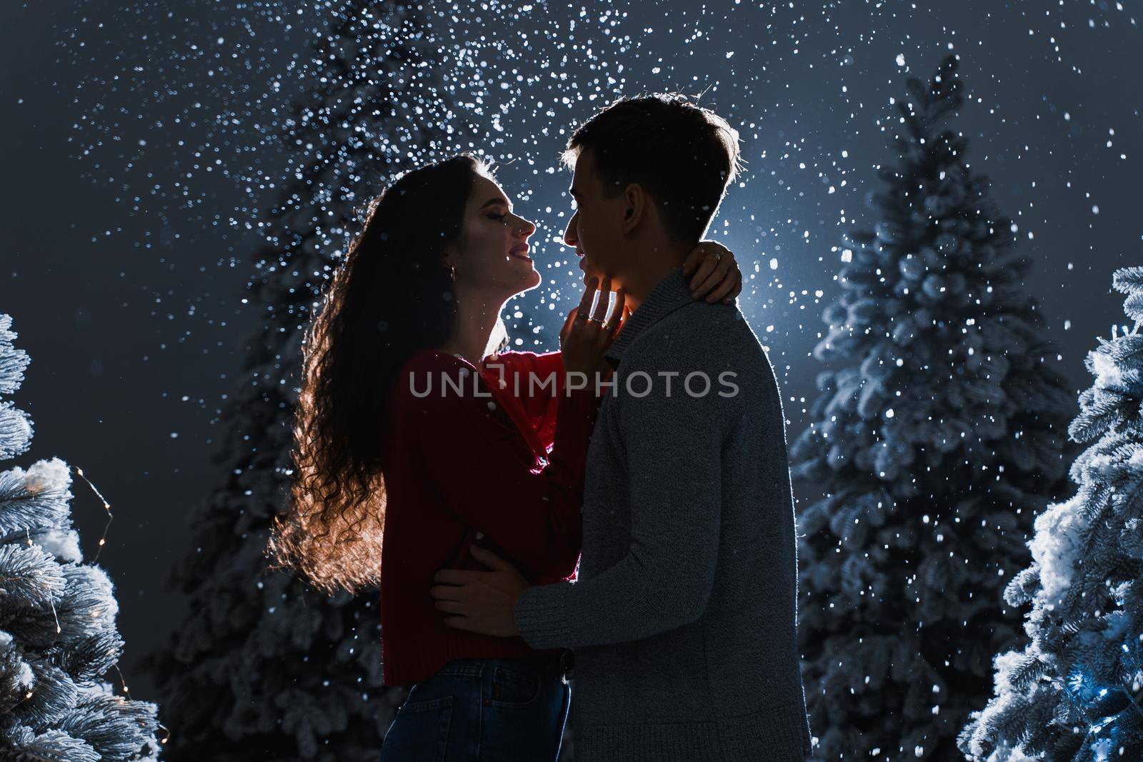 Falling snow and kisses with falling snow on dark blue background. Happy young couple hug and kiss near christmas trees at the eve of new year celebration in winter day