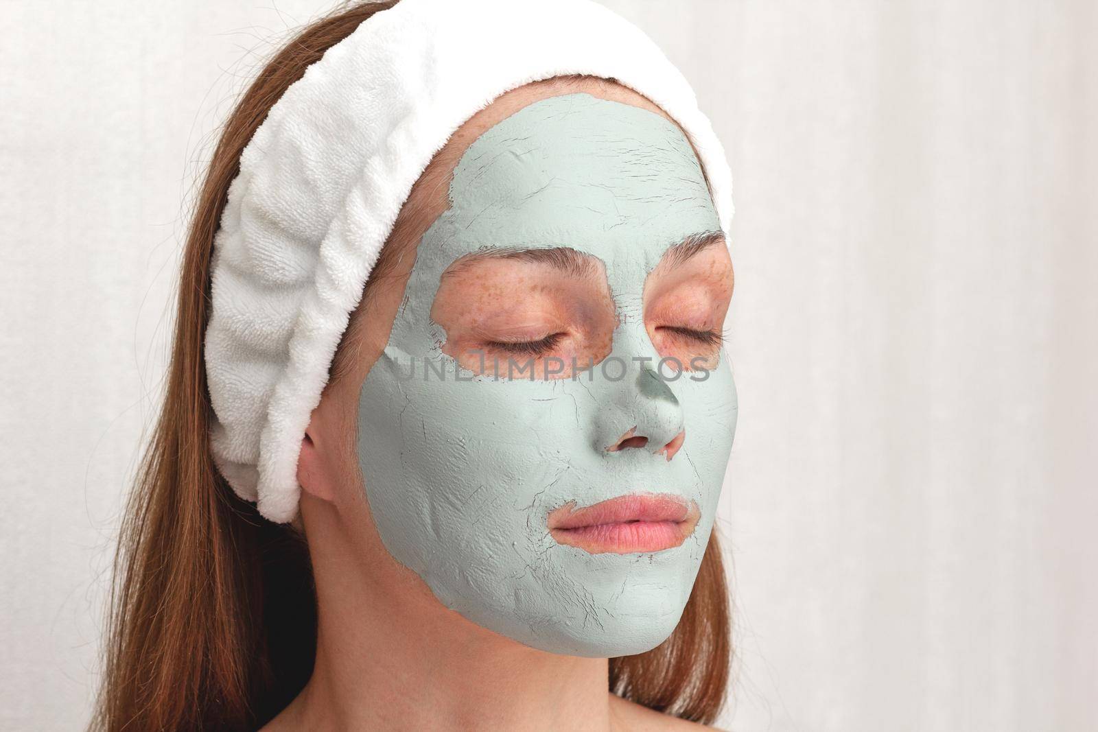 Woman with white band on head with blue dry clay mask on face with closed eyes over white background