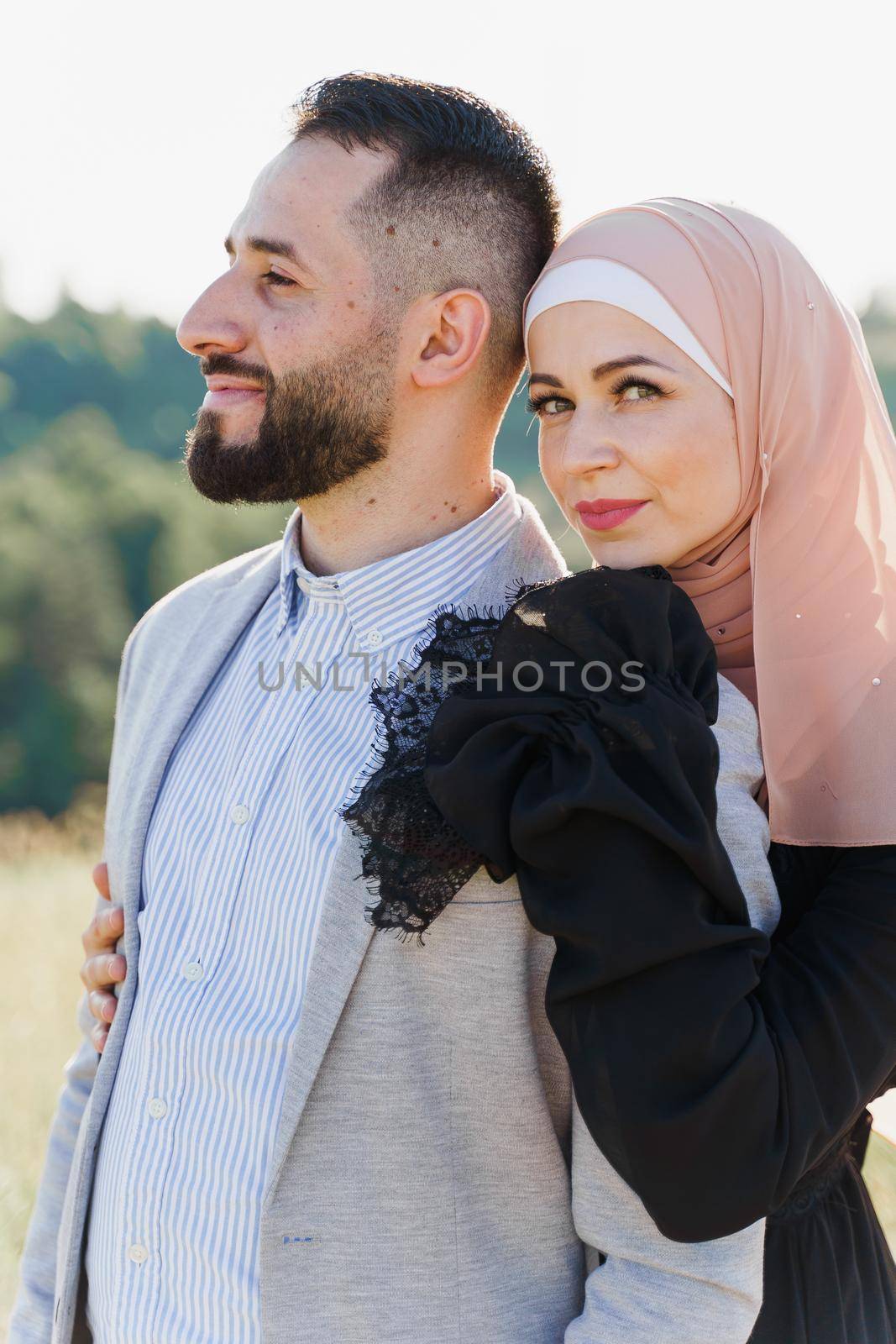 Muslim love story close-up. Mixed couple smiles and hugs on the green hills . Woman weared in hijab looks to her man. Advert for on-line dating agency