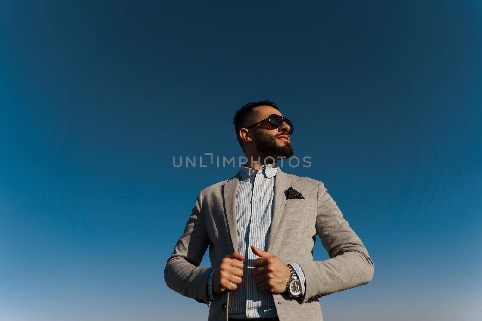 Handsome bearded arabian man in sunglasses on the blue sky background. Confident muslim business man. Arab man weared in grey coat. by Rabizo