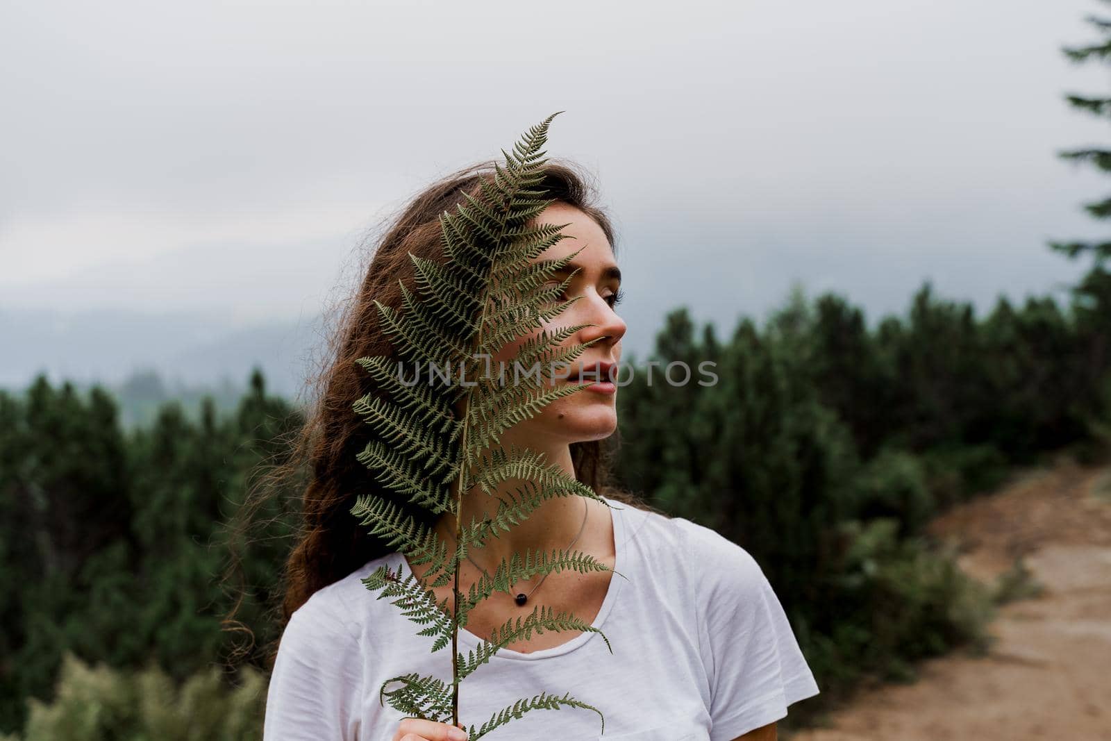 Girl is holding leaf of fern and travelling in the mountains. Foggy day. Hiking and climbing up to the peak of the mountain.