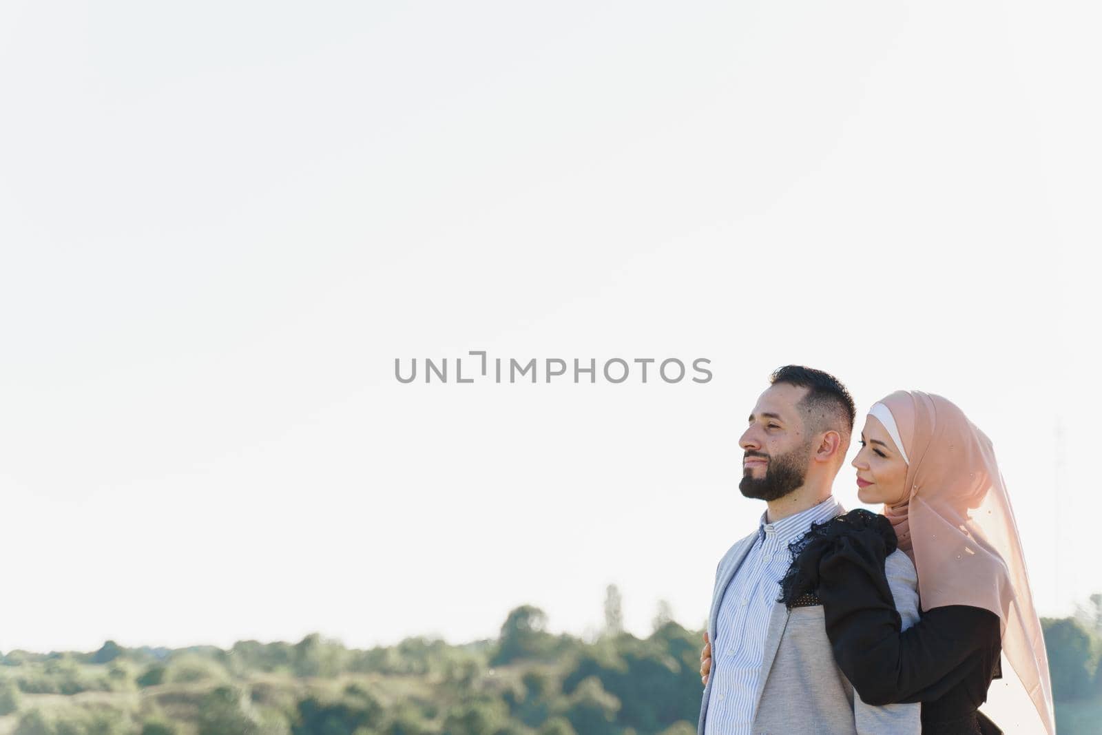 Muslim love story. Mixed couple smiles and hugs on the green hills . Woman weared in hijab looks to her man. Advert for on-line dating agency. by Rabizo