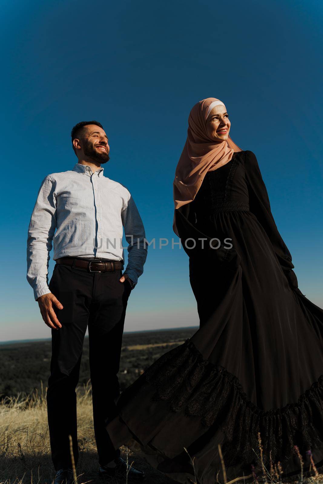 Muslim love story on the blue sky background. cheerful mixed couple smiles and hugs . Woman weared in hijab looks to her man. Advert for on-line dating agency. by Rabizo