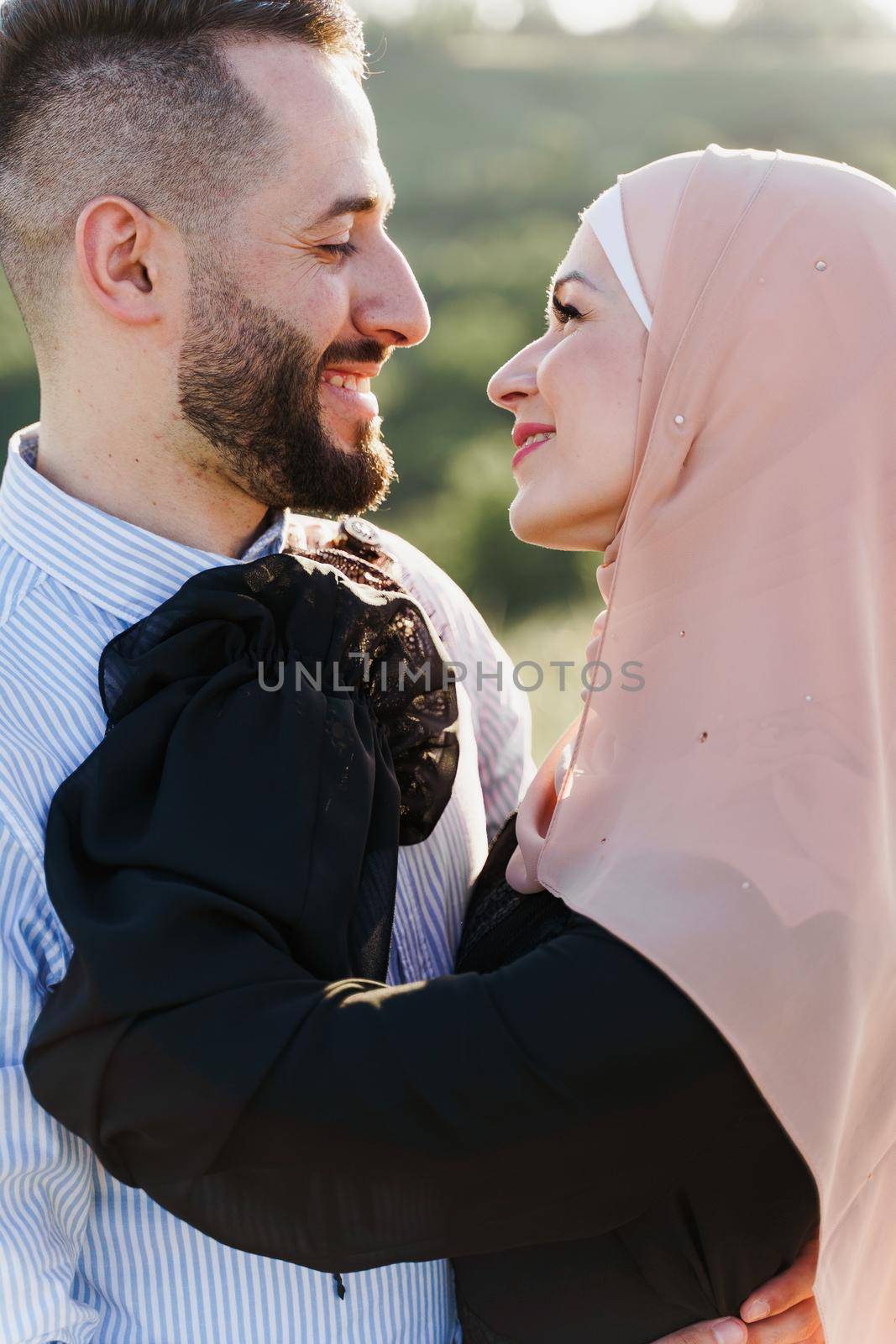 Muslim love story close-up. Mixed couple smiles and hugs on the green hills . Woman weared in hijab looks to her man. Advert for on-line dating agency