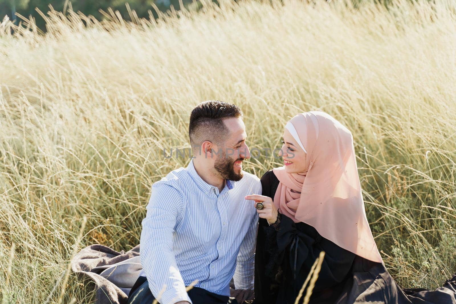 Muslim love story. Mixed couple seats on the grass, smiles and hugs.. Woman weared in hijab looks to her man. Advert for on-line dating agency. by Rabizo