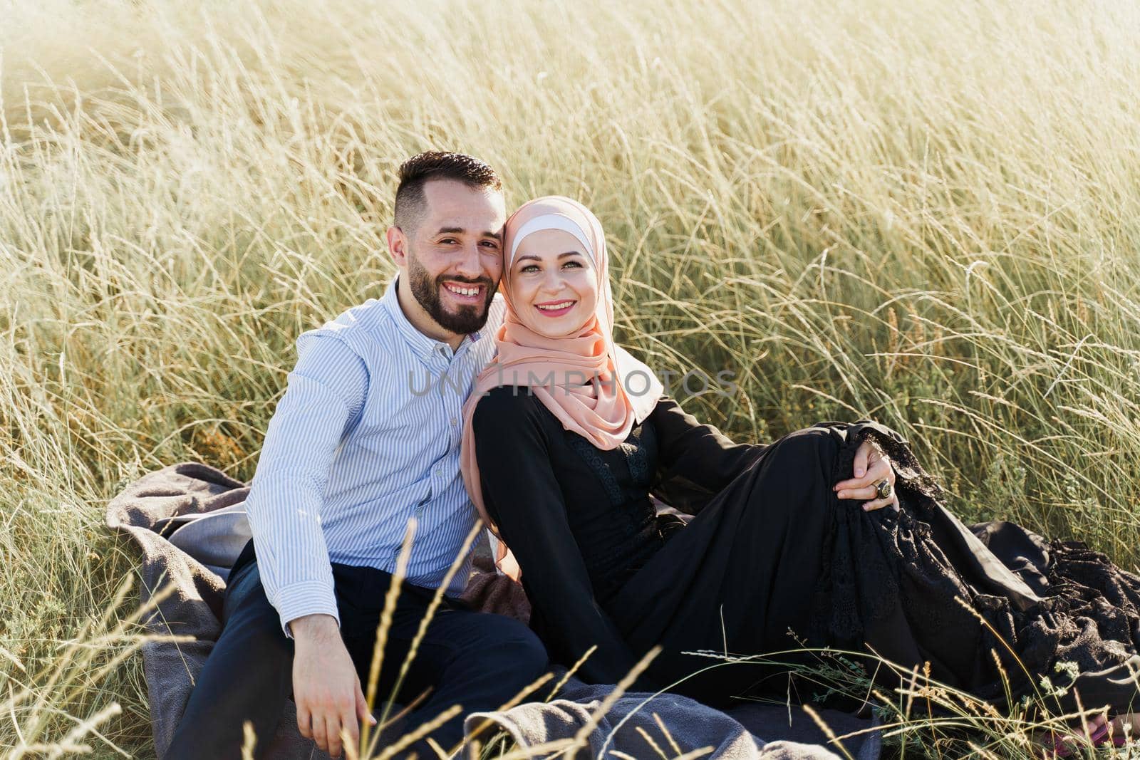 Muslim love story. Mixed couple seats on the grass, smiles and hugs.. Woman weared in hijab looks to her man. Advert for on-line dating agency