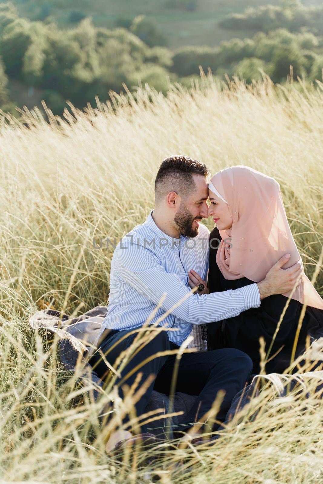 Muslim love story. Mixed couple seats on the grass, smiles and hugs.. Woman weared in hijab looks to her man. Advert for on-line dating agency. by Rabizo