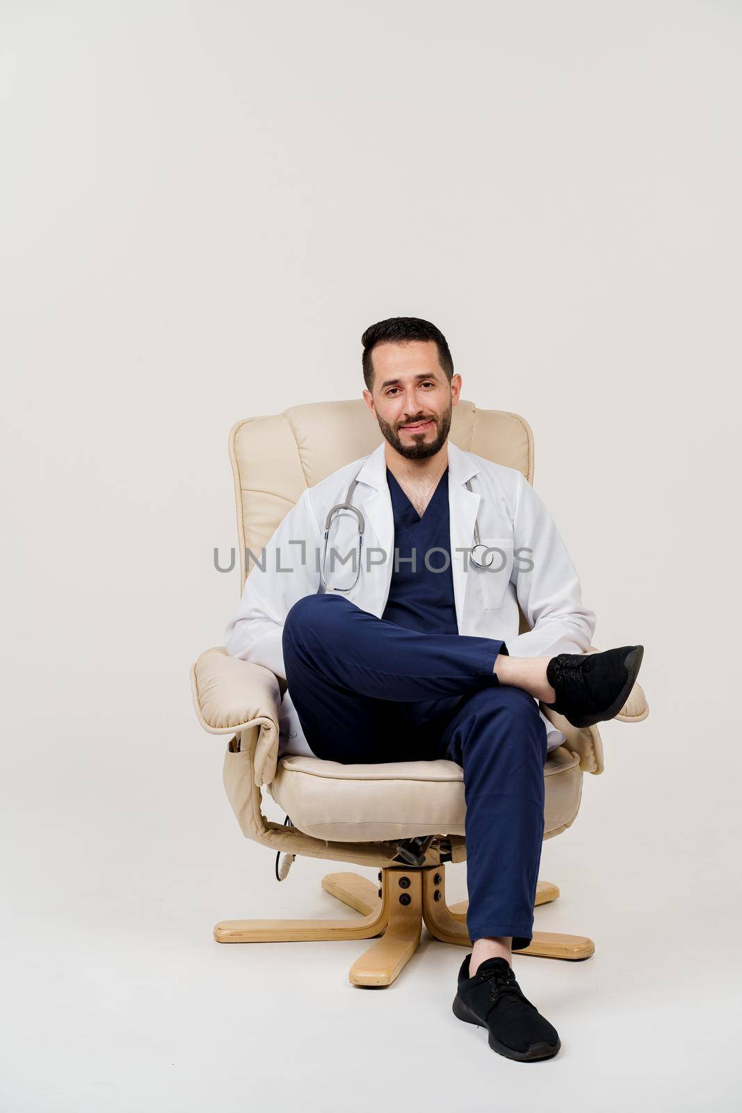 Arabian doctor surgeon in medical robe with stethoscope seats in armchair in studio on white blanked background. Handsome arab on white background