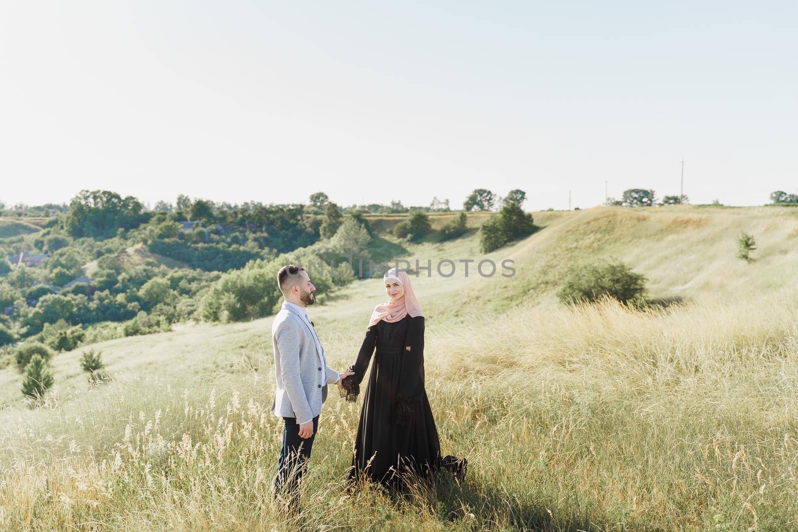 Muslim love story of mixed couple. Man and woman smiles and walks on the green hills . Woman weared in hijab looks to her man. Advert for on-line dating agency