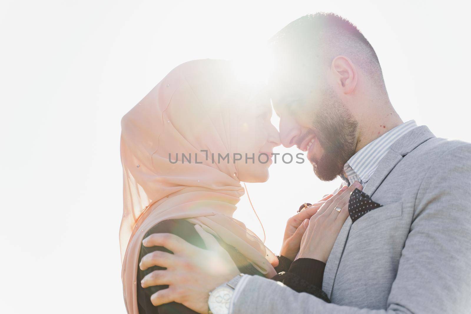 Muslim love story close-up with sun light. Mixed couple smiles and hugs at sunset. Woman weared in hijab looks to her man. Advert for on-line dating agency