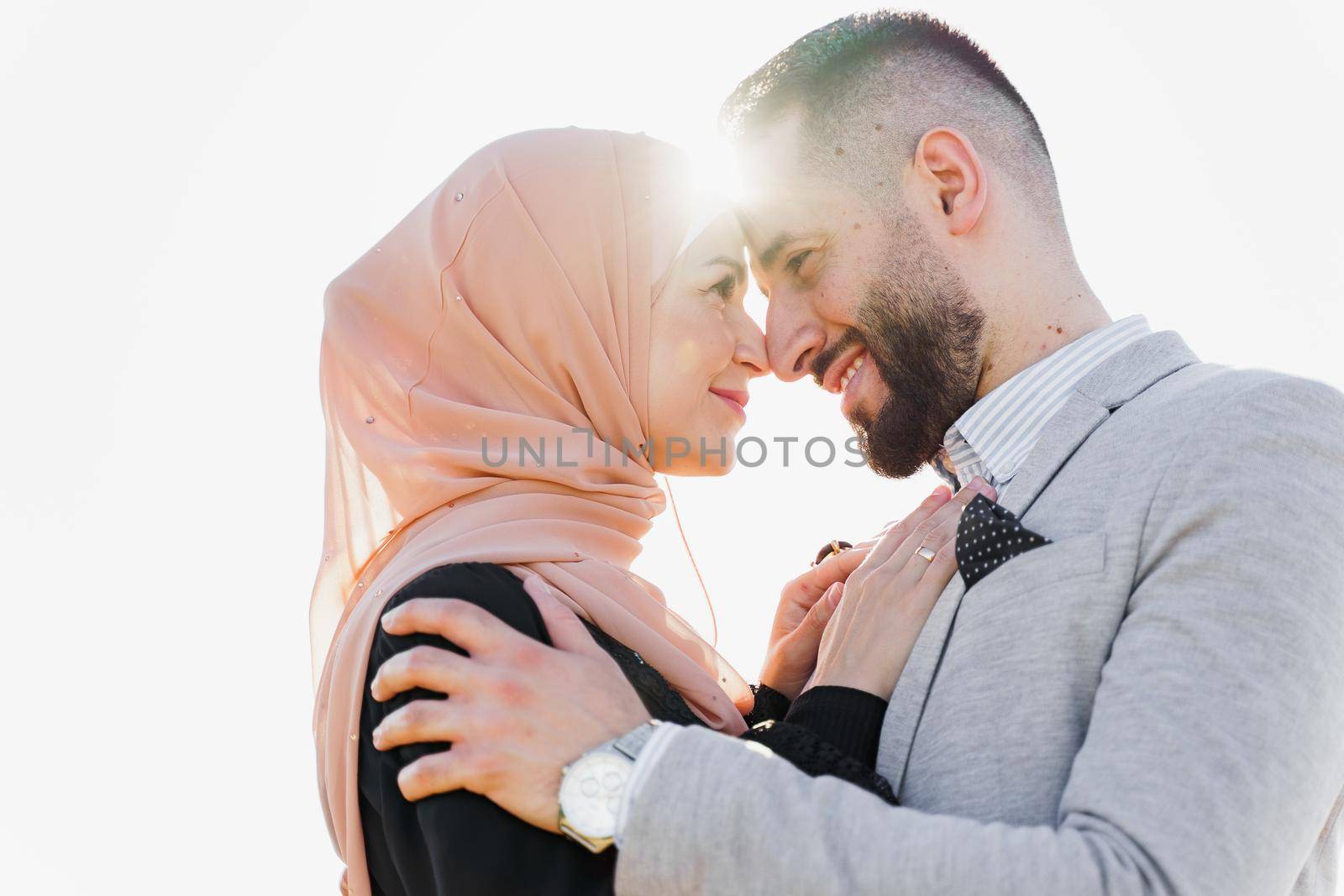 Muslim love story close-up with sun light. Mixed couple smiles and hugs at sunset. Woman weared in hijab looks to her man. Advert for on-line dating agency