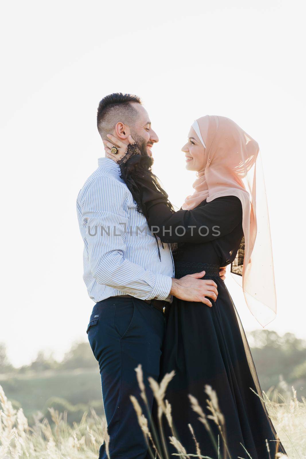 Muslim love story with sun light. Mixed couple smiles and hugs at sunset. Woman weared in hijab looks to her man. Advert for on-line dating agency