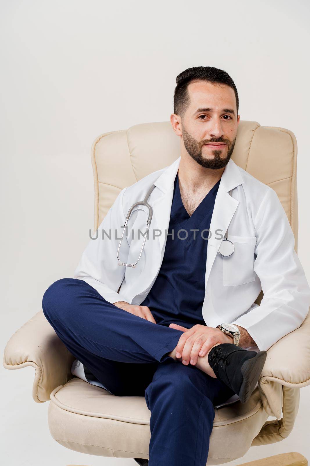 Arabian doctor surgeon in medical robe with stethoscope seats in armchair in studio on white blanked background. Handsome arab on white background