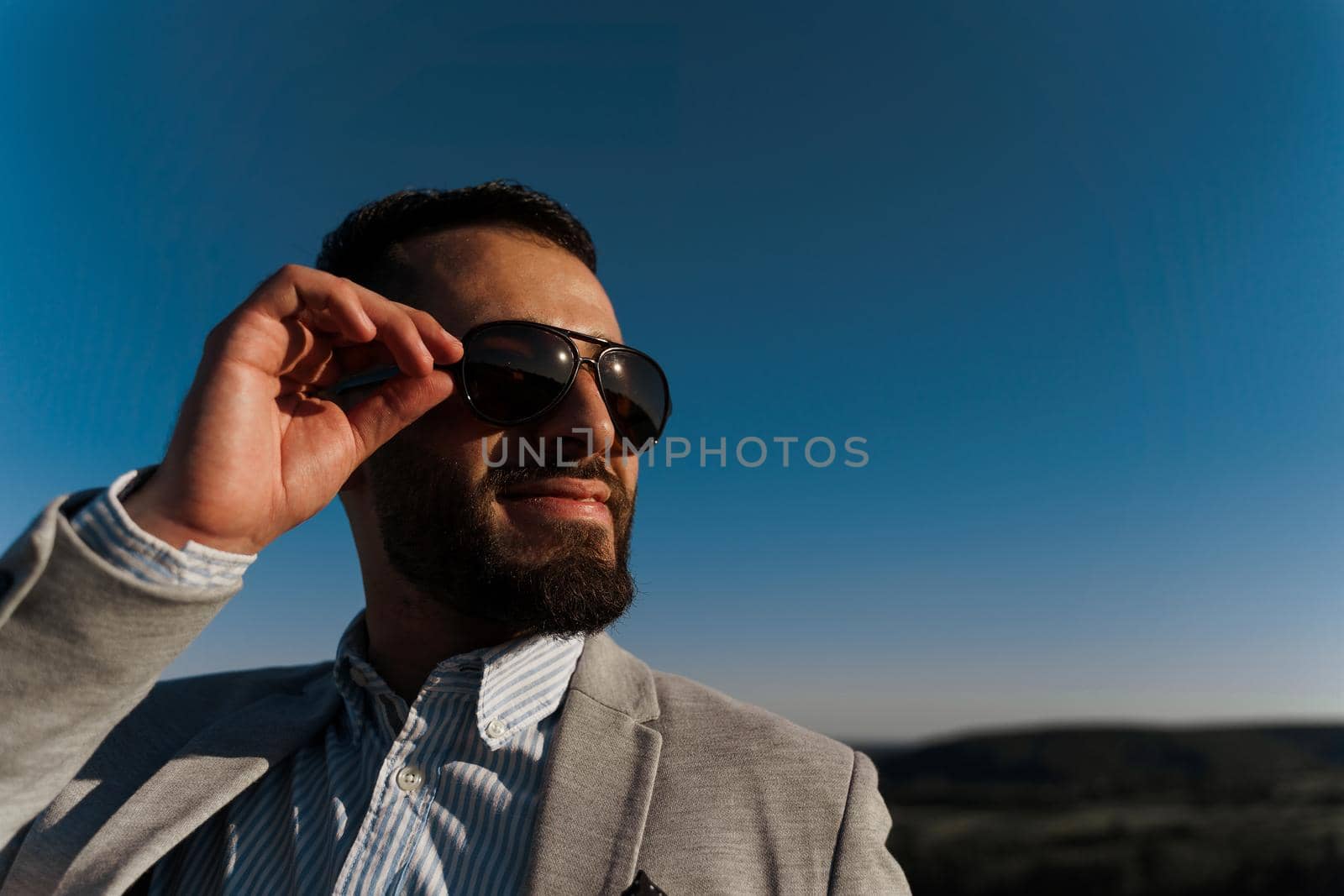 Handsome bearded arabian man in sunglasses on the blue sky background. Confident muslim business man. Arab man weared in grey coat. by Rabizo