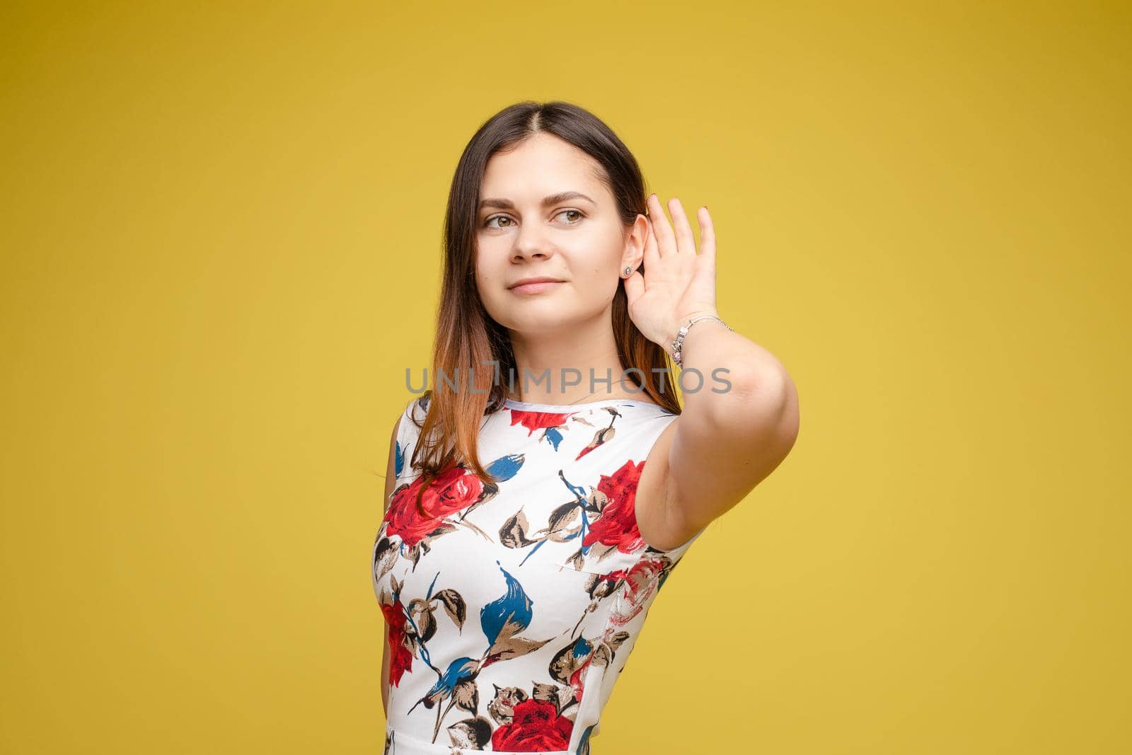 Close up of pretty young lady holding her hand near ear and listening carefully. Beautiful brunette woman posing at camera and smiling. Happy attractive woman in dress with red flowers hearing noises.