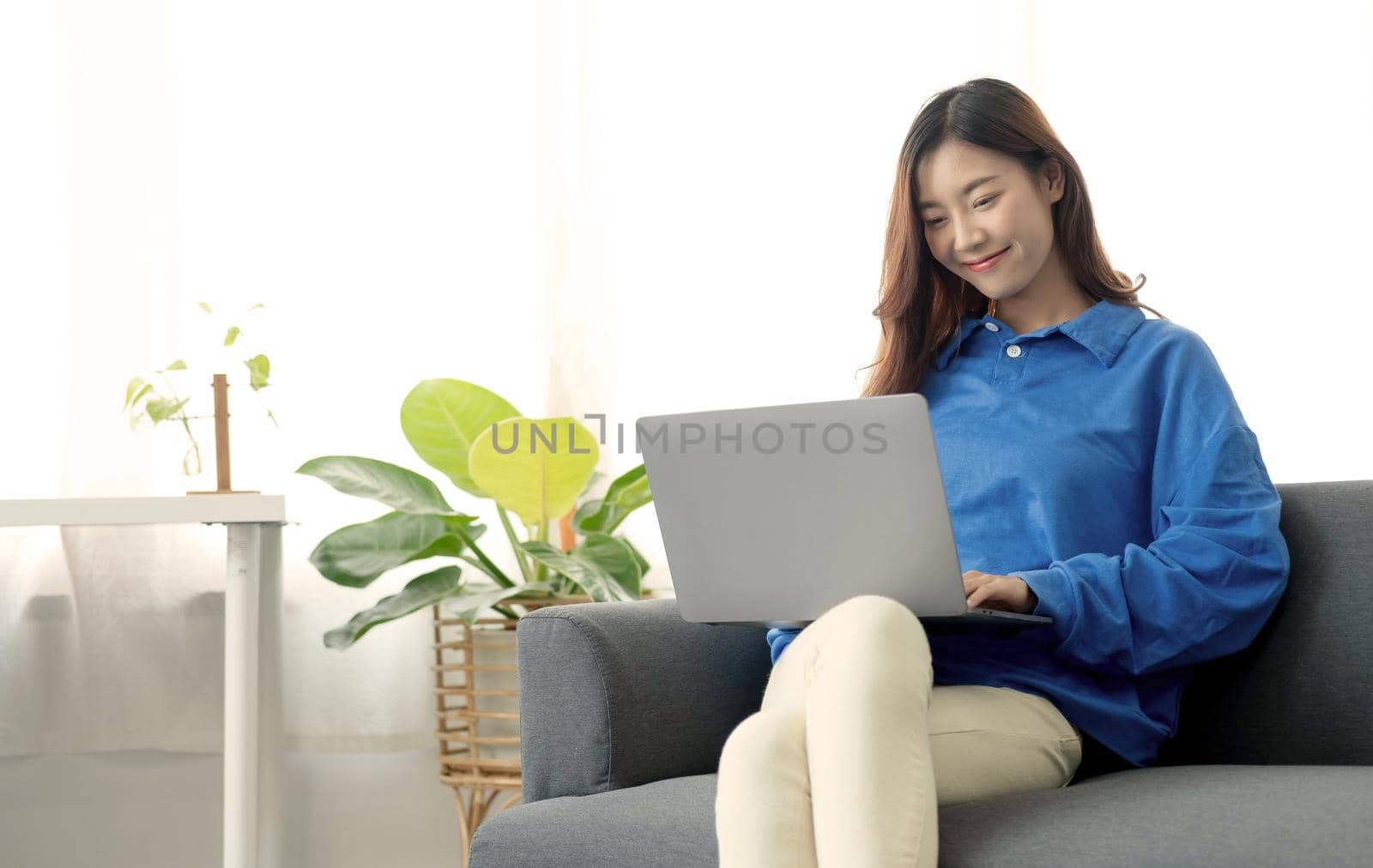 Young business freelance Asian woman working on laptop checking social media while lying on the sofa when relax in living room at home. by wichayada