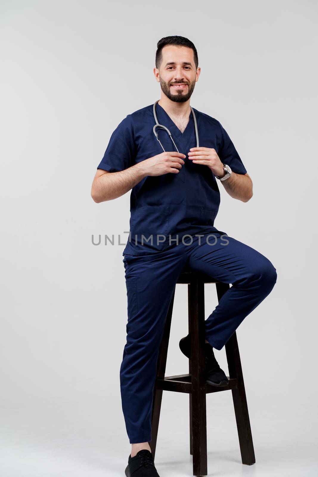 Handsome arab doctor surgeon smiles on white background. Bearded student with stethoscope in medical robe seats on chair on blanked background.