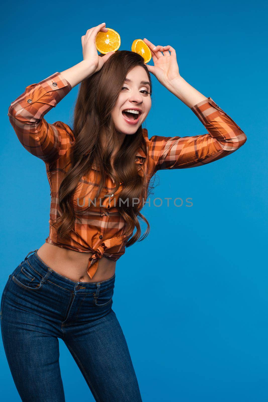 Young girl in casual clothes hiding her face behind fruits. Brunette lady in checkered shirt and jeans holding halves of oranges as sunglasses. Beatiful cute woman with opened belly happily smiling.