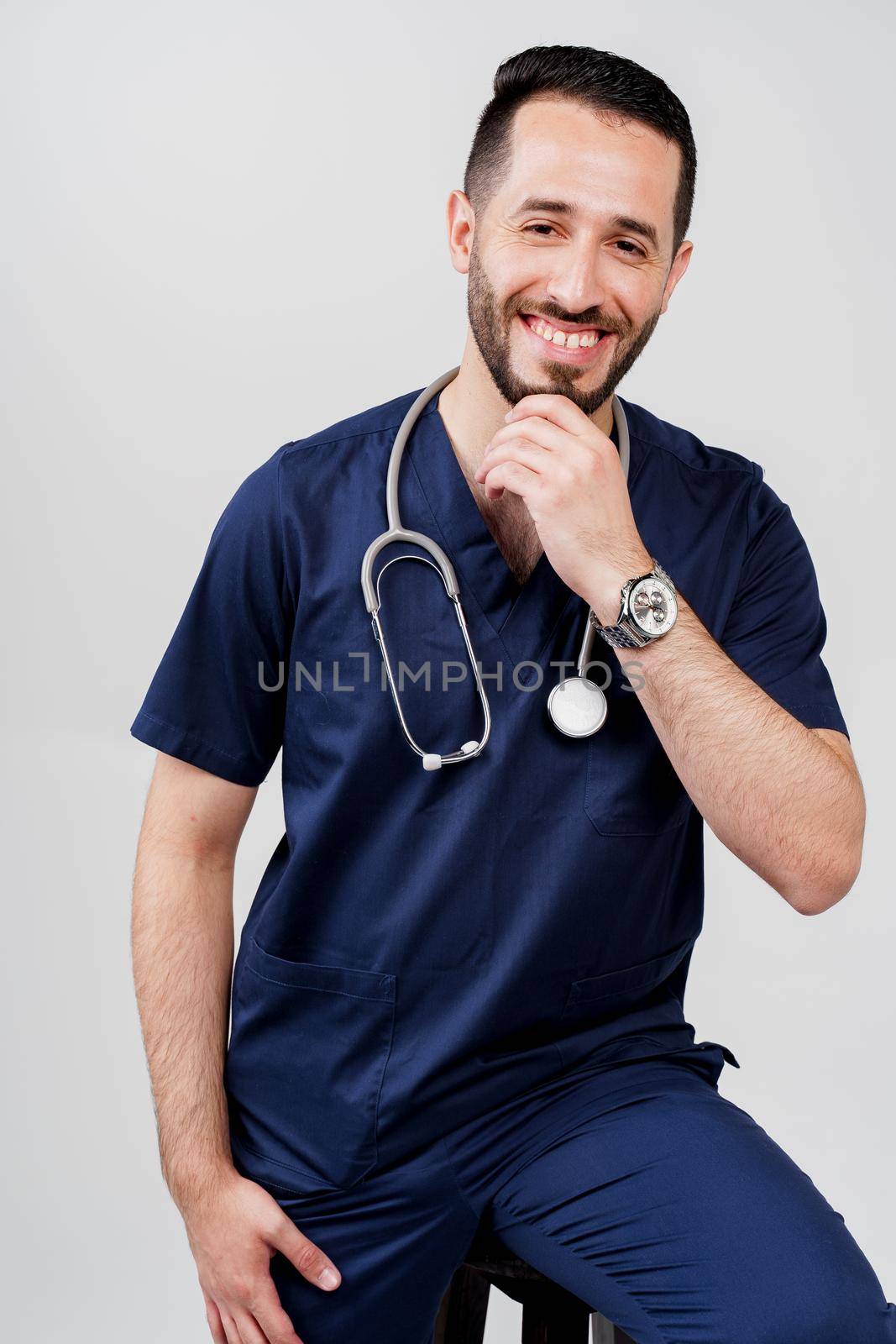 Arabian medical student with stethoscope in surgical uniform smiles on blanked background. Handsome bearded man in studio. Confident doctor at consultation