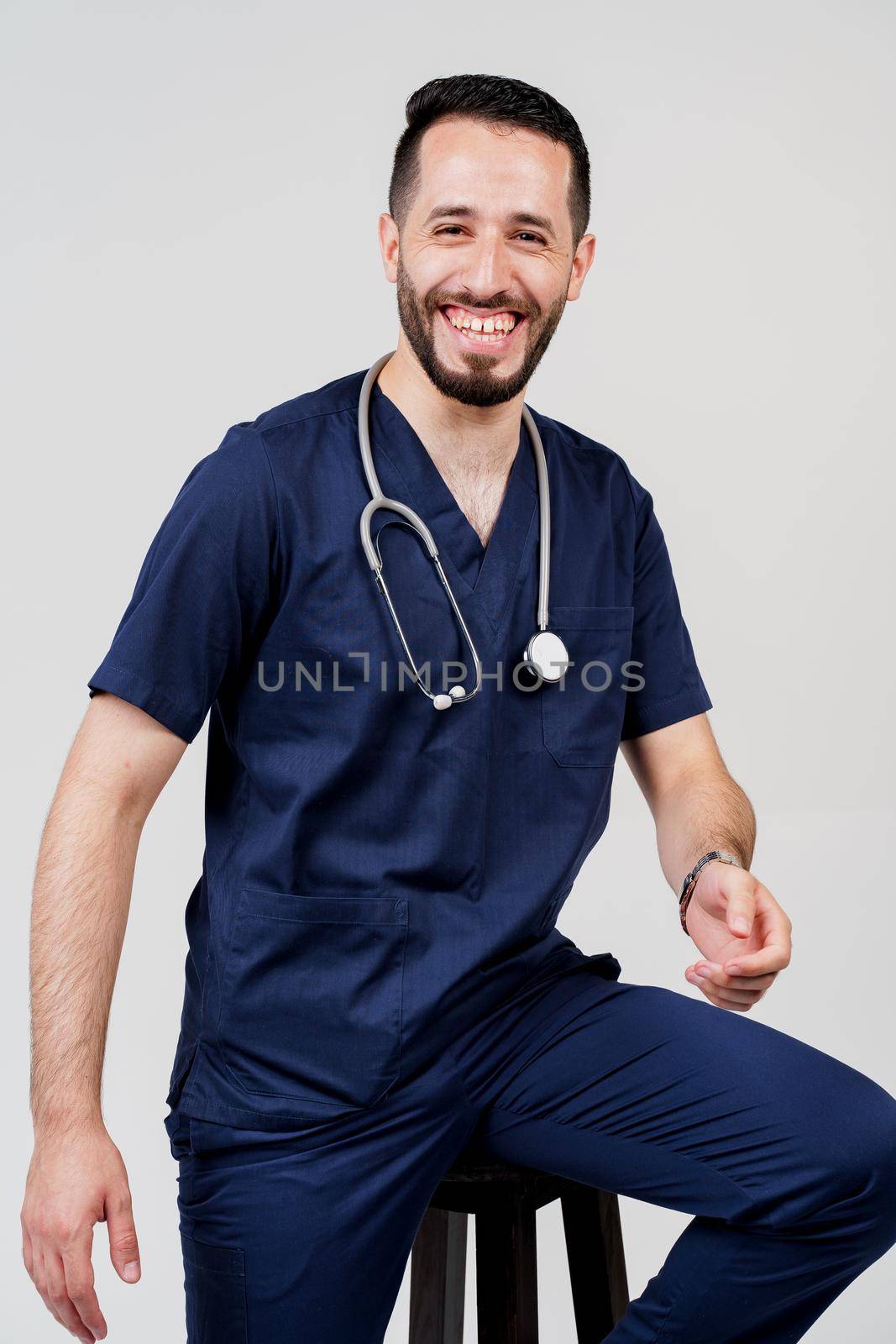 Arabian medical student with stethoscope in surgical uniform smiles on blanked background. Handsome bearded man in studio. Confident doctor at consultation. by Rabizo