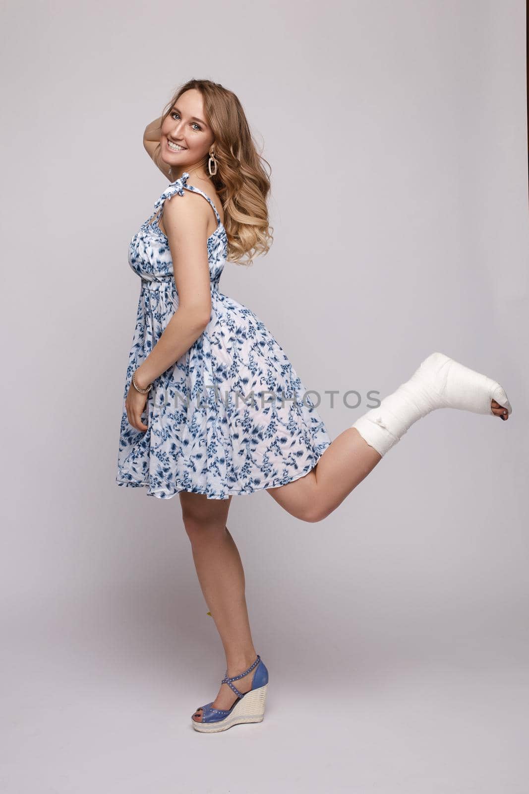 Side view of pretty woman in short dress posing with broked leg on isolated background. Happy confident model with fracture looking at camera and laughing. Concept of happiness and inquiry.
