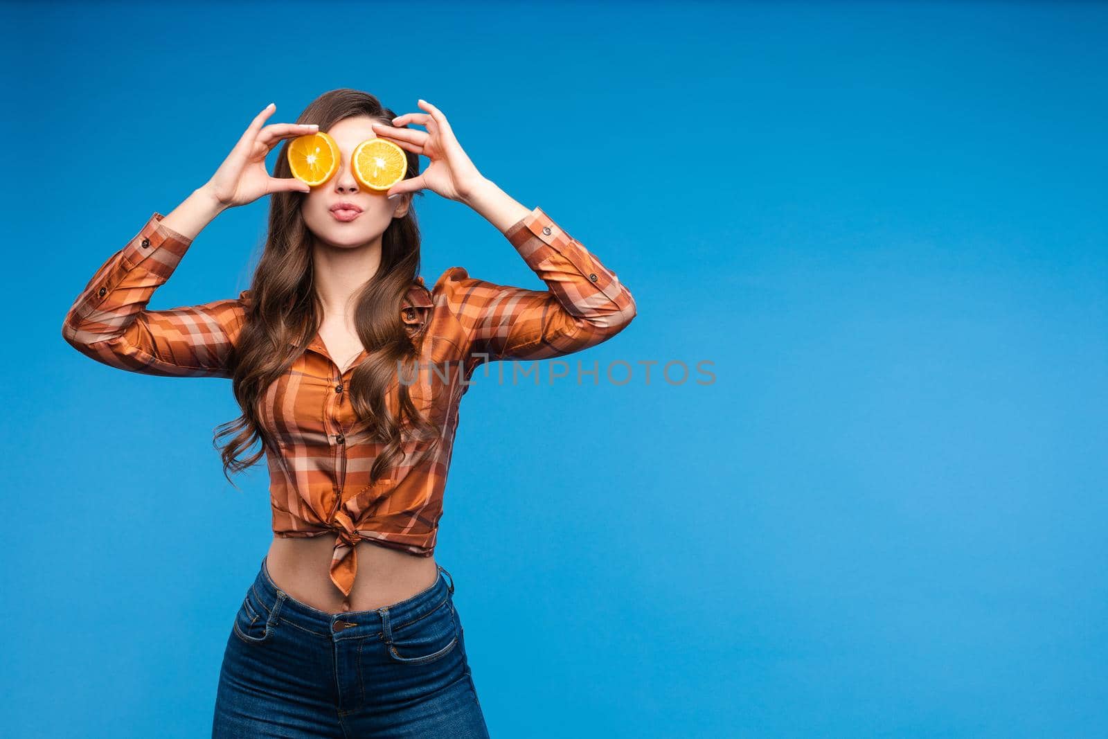 Young girl in casual clothes hiding her face behind fruits. Brunette lady in checkered shirt and jeans holding halves of oranges as sunglasses. Beatiful cute woman with opened belly happily smiling.