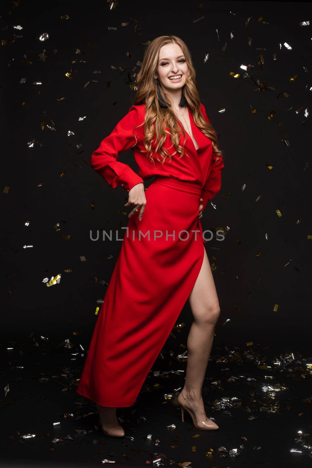 Happy young girl looking surprisingly while golden confetti falling. Beautiful woman in emerald elegant long dress smiling and dancing among decorations. Gorgeous lady celebrating birthday on party.