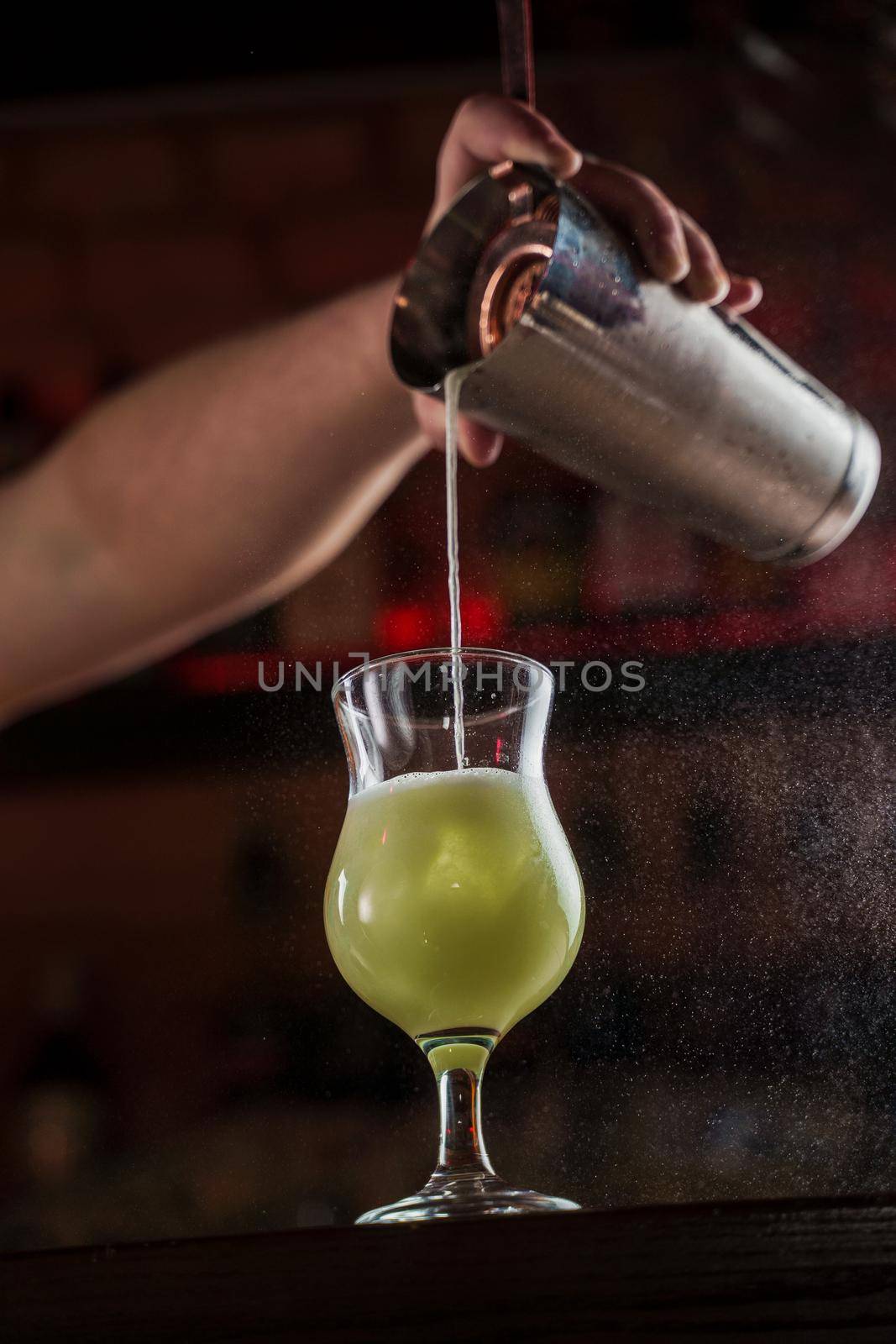 Bartender sprinkles on illuminated glass with bright green cold cocktail on bar counter and makes fire flame over it by Rabizo